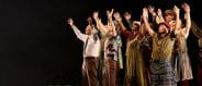 A group of people dressed in earth tones stand with hands raised aloft