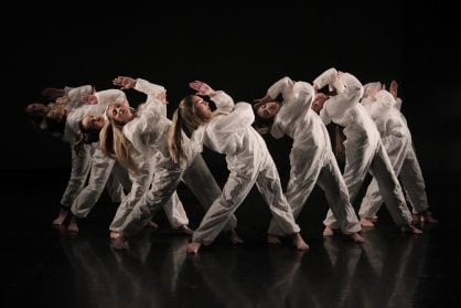 A group of five+ girls dressed in white paper suits dance together in formation bending to the side