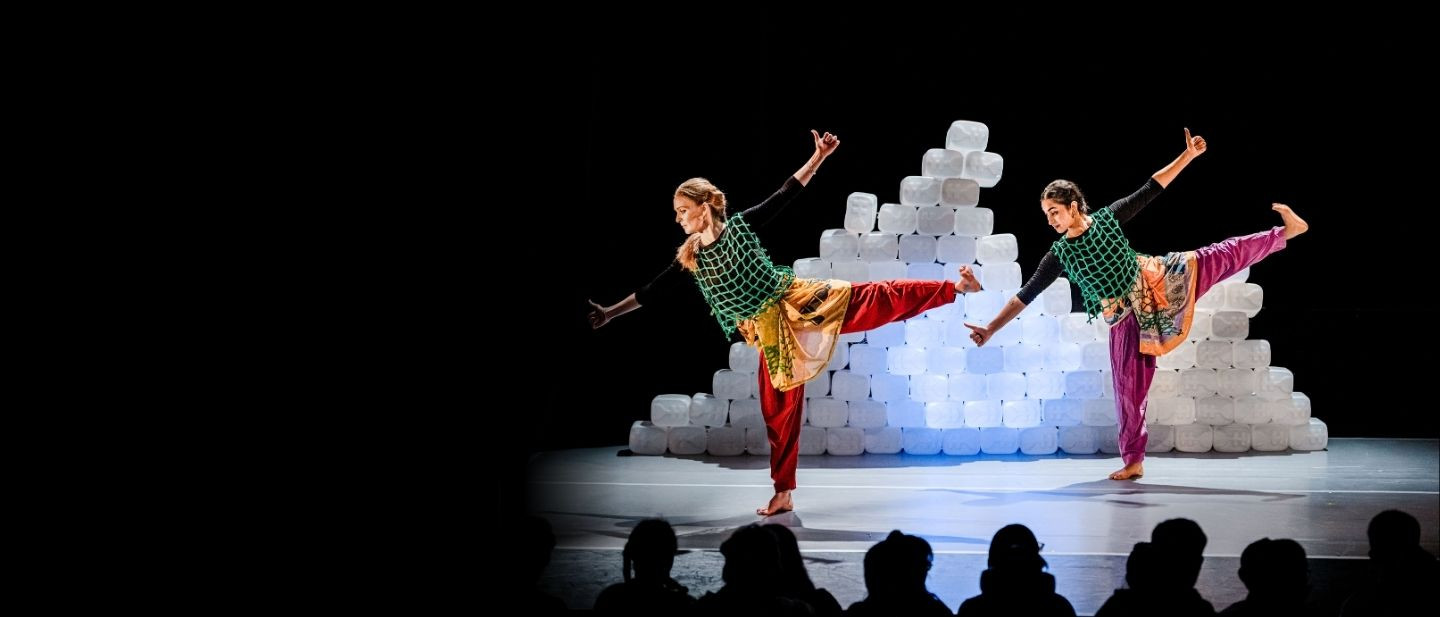 Two dancers balance on one leg with their arms and legs outstretched. They wear costumes made from discarded plastic.