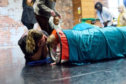 A woman crouches at the end of a play tunnel covering her eyes as a toddler peeks out the end. 