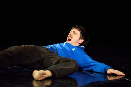 A performer lies on the stage floor with their head raised. They look as though they are shouting.