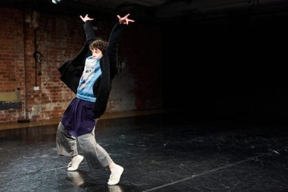 A performer is on a stage with lots of layers of clothing on. they have both arms in the air 