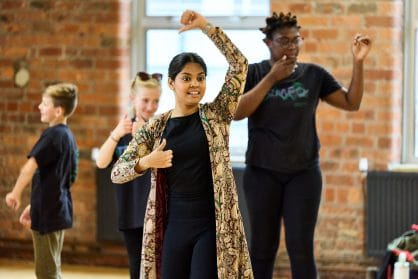 A dancer is leading some children. She is smiling with one arm in front of her and the other in the air.