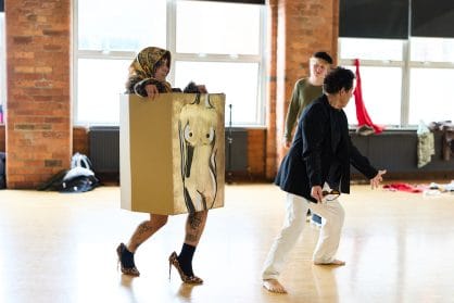 A person walks wearing heels and a large cardboard box with a woman's torso sketched across the front