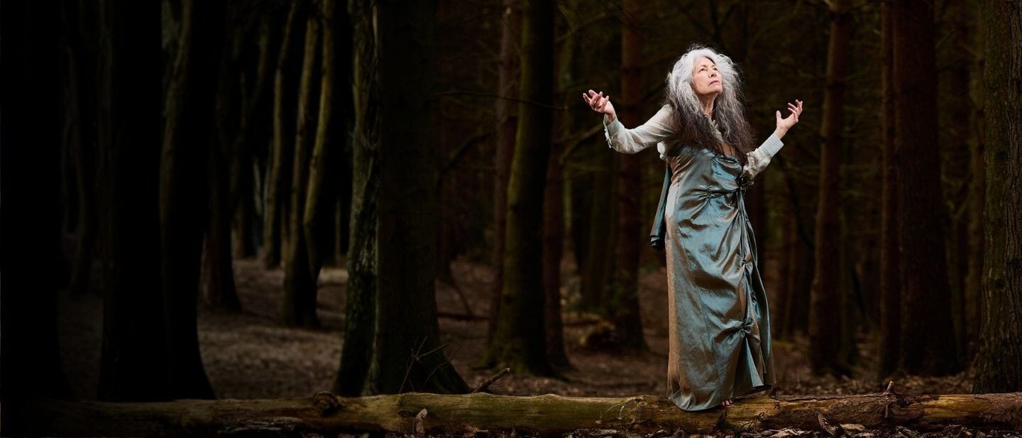 A woman in silk robes stands in a wood with arms in the air