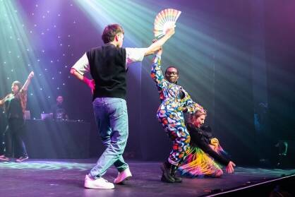 Two people strut on a stage with a fan