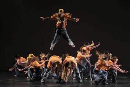 A group of street dancers dressed in yellow are crouched while one dancer jumps high in the air