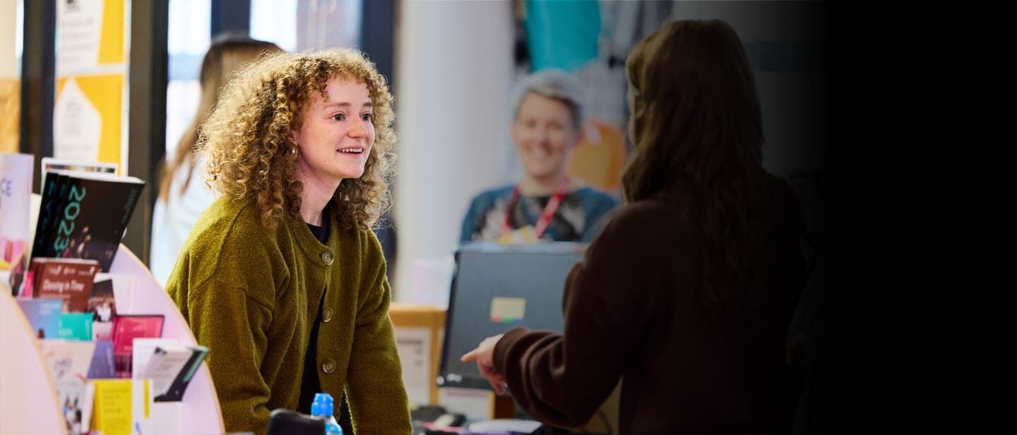 A front of house staff member leans over the reception desk to help someone