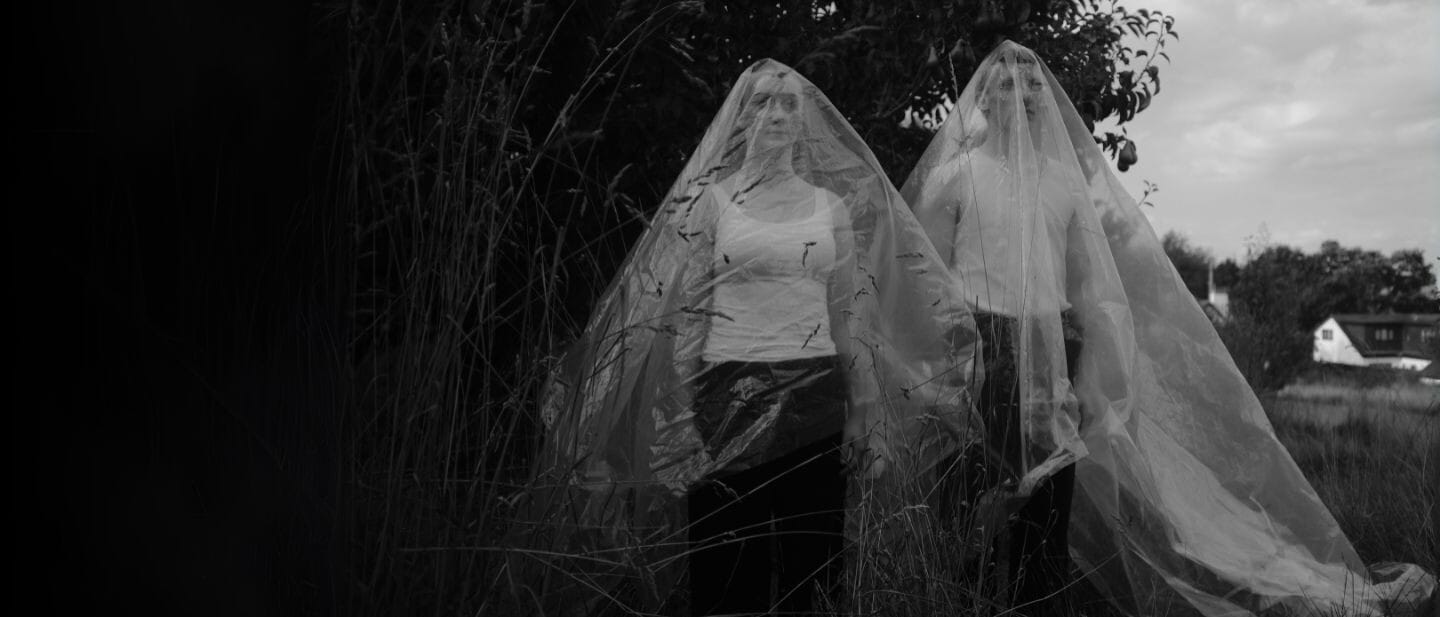2 people stand in nature with serious faces, they have a transparent plastic draped over them